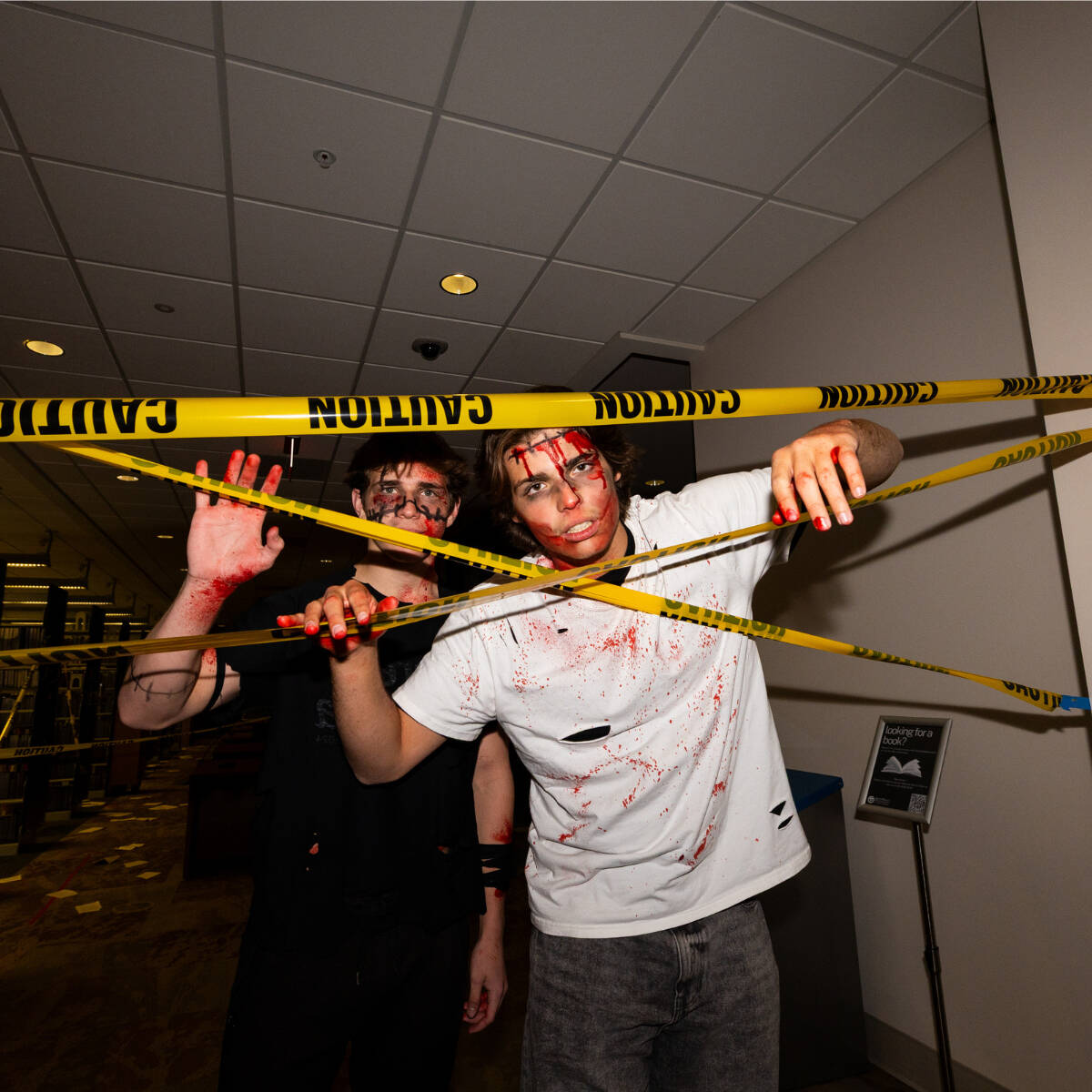 Two students dressed as zombies pushing through yellow caution tape in the library